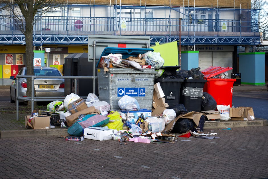 An overflowing collection of mixed waste and rubbish, including crumpled cardboard boxes, plastic and paper bags, discarded packaging, and miscellaneous debris, sit piled around a large gray recycling bin marked for mixed paper and card. The waste extends onto the sidewalk, with some items spilling over the edges of the bins, indicating a significant accumulation of refuse in an urban environment. Behind the rubbish, there are parked cars on the street, including a silver vehicle, and a building with storefronts and signage, partially obscured by blue scaffolding and a tree with bare branches. The scene is set during daylight under natural lighting, and the image captures the surrounding urban setting distinctively. This type of scene illustrates the importance of independent rubbish collection services like those offered by Waste Removal Balham, emphasizing the need for effective and responsible waste management outside conventional municipal services.