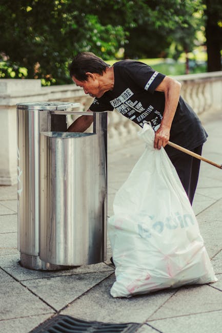 An overflowing collection of mixed waste and rubbish, including crumpled cardboard boxes, plastic and paper bags, discarded packaging, and miscellaneous debris, sit piled around a large gray recycling bin marked for mixed paper and card. The waste extends onto the sidewalk, with some items spilling over the edges of the bins, indicating a significant accumulation of refuse in an urban environment. Behind the rubbish, there are parked cars on the street, including a silver vehicle, and a building with storefronts and signage, partially obscured by blue scaffolding and a tree with bare branches. The scene is set during daylight under natural lighting, and the image captures the surrounding urban setting distinctively. This type of scene illustrates the importance of independent rubbish collection services like those offered by Waste Removal Balham, emphasizing the need for effective and responsible waste management outside conventional municipal services.