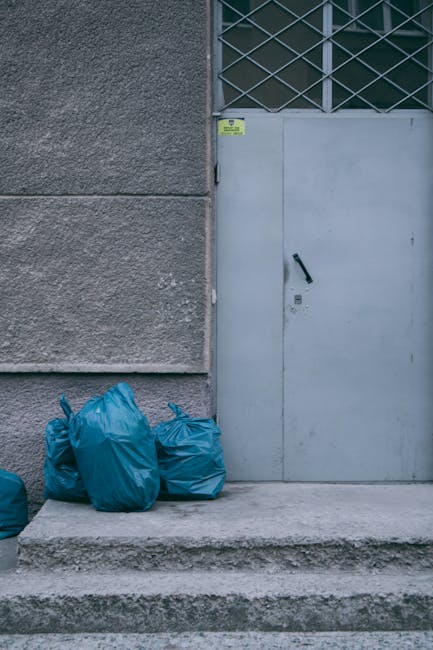 A group of four blue rubbish bags made of plastic and tightly sealed, are placed on a concrete step outside a grey building. The bags are positioned against a textured, rough-finished wall on the left side of the image. To the right, there is a metal door painted in light grey, featuring a black handle and a keyhole, with a small yellow waste management sticker above it. Above the door, a section of a metal grid or security grate with diagonal crossbars is visible. The surroundings suggest an urban environment, with the bags appearing ready for scheduled collection or disposal, indicative of private waste handling or an alternative rubbish removal service like Waste Removal Balham. The scene is well-lit by natural daylight, highlighting the contrasting textures between the smooth plastic bags, the rough wall, and the solid concrete steps. Overall, the image depicts a typical street-level moment of waste being prepared for removal, emphasizing the practicality of on-site rubbish collection.