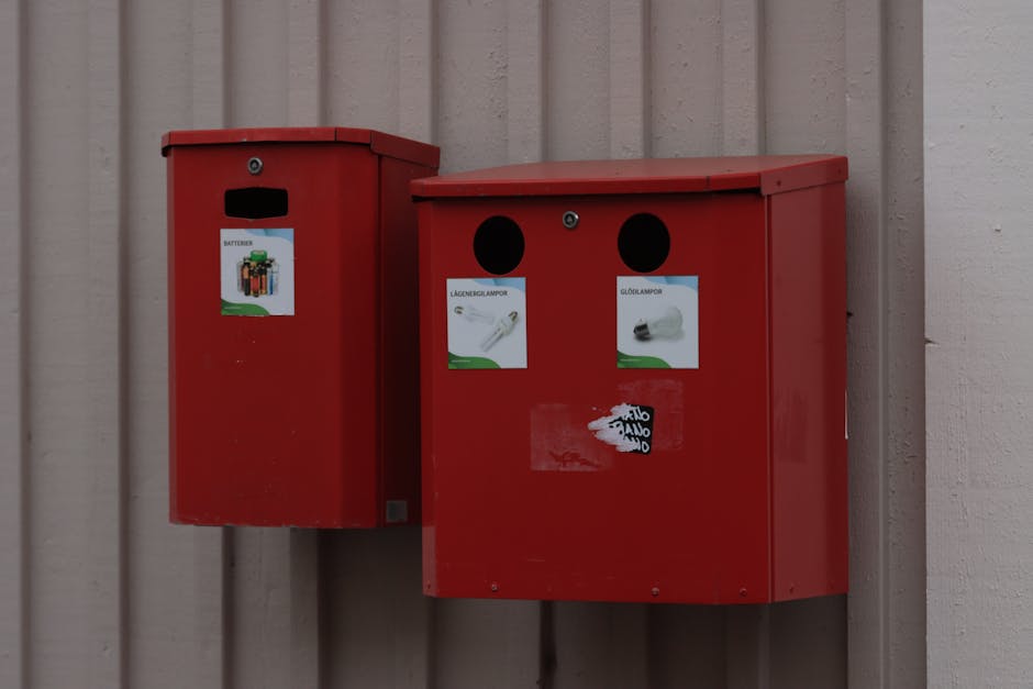 Two red utility boxes mounted on a beige wooden wall, used for waste or recycling collection. The left box is taller with a small rectangular slot near the top and a sticker depicting a set of batteries, indicating it is for electronic waste. The right, slightly wider box has two circular openings at the top, and each has a printed label beneath it; one label shows a light bulb labeled 'Lagergrenlampor' (storage lamps) in Scandinavian language, and the other displays a similar label for a bulb with a light-colored image. Both boxes have a metallic finish, with the larger one featuring a transparent plastic sticker below the labels. The surrounding environment suggests an outdoor setting, with natural light illuminating the scene, and the boxes positioned close to the wall, indicating a designated collection point for specific waste materials. These structures may be part of an independent waste collection system, supporting private disposal and alternative rubbish management options, with the branding 'Waste Removal Balham' subtly present in the context of rubbish services.