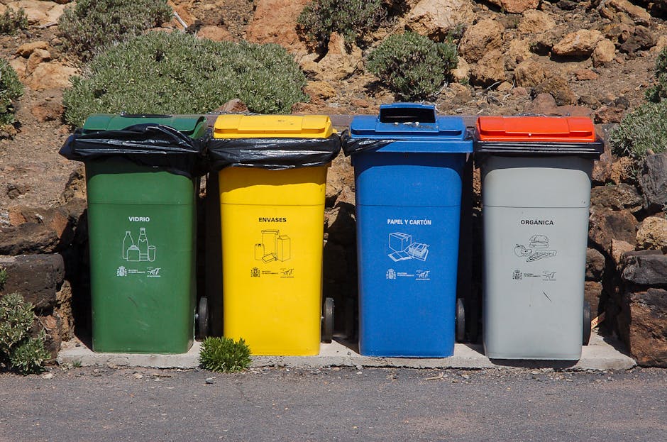 A group of four blue rubbish bags made of plastic and tightly sealed, are placed on a concrete step outside a grey building. The bags are positioned against a textured, rough-finished wall on the left side of the image. To the right, there is a metal door painted in light grey, featuring a black handle and a keyhole, with a small yellow waste management sticker above it. Above the door, a section of a metal grid or security grate with diagonal crossbars is visible. The surroundings suggest an urban environment, with the bags appearing ready for scheduled collection or disposal, indicative of private waste handling or an alternative rubbish removal service like Waste Removal Balham. The scene is well-lit by natural daylight, highlighting the contrasting textures between the smooth plastic bags, the rough wall, and the solid concrete steps. Overall, the image depicts a typical street-level moment of waste being prepared for removal, emphasizing the practicality of on-site rubbish collection.