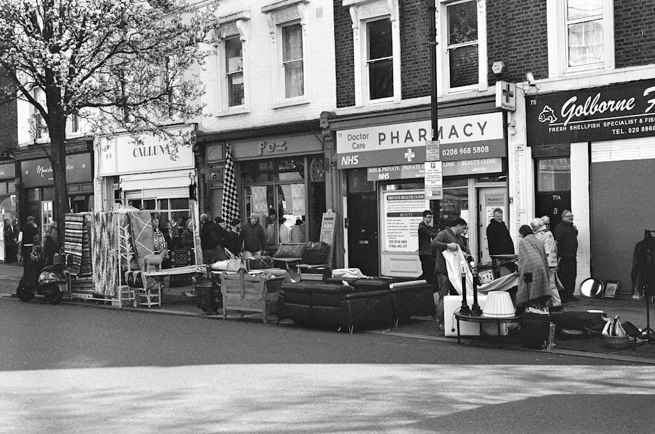 The image shows a busy street scene with various second-hand or discarded furniture and household items arranged along the pavement in front of a row of commercial buildings, including a pharmacy and a fish and seafood shop. There are sofas with visible fabric upholstery, tables, lamps, and other miscellaneous furniture items placed close together, some overlapping slightly. The furniture appears used, with some items showing signs of wear. Several people are gathered around the pile, some engaging in conversation or inspecting the items, while others stand nearby, possibly observing or waiting. A small scooter is parked to the left, along with a few other pedestrians in the background. The storefronts have signage, with one indicating a pharmacy and another a seafood business. The scene is lit by natural daylight, with a tree visible on the left side of the image, and the overall environment suggests an informal or on-site clearance situation, possibly related to local waste disposal or a community clearance, with Waste Removal Balham likely involved in the removal or collection of these items. The overall composition and context reflect typical roadside furniture and household item collection aimed at reuse or alternative waste handling.