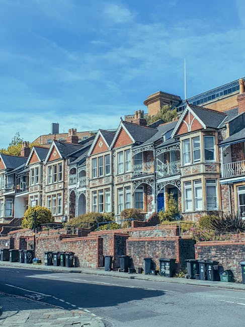 A row of Victorian-style terraced houses with decorative wrought iron balconies, bay windows, and intricate brickwork detail, situated on a slight incline above a paved street. The front gardens are enclosed by low red brick walls, with neatly trimmed bushes and small trees visible along the fence line. Multiple black wheelie bins are placed along the edge of the pavement, indicating waste collection points. The background features modern buildings with glass facades, and the sky above is bright blue with some scattered clouds, suggesting clear weather. This scene highlights a residential area where private rubbish disposal may be arranged for property clearance or regular waste removal, aligning with independent waste management services. Waste Removal Balham's presence is implied through the context of rubbish handling in this urban setting, showcasing typical features of a quiet, well-maintained neighbourhood during daylight hours.