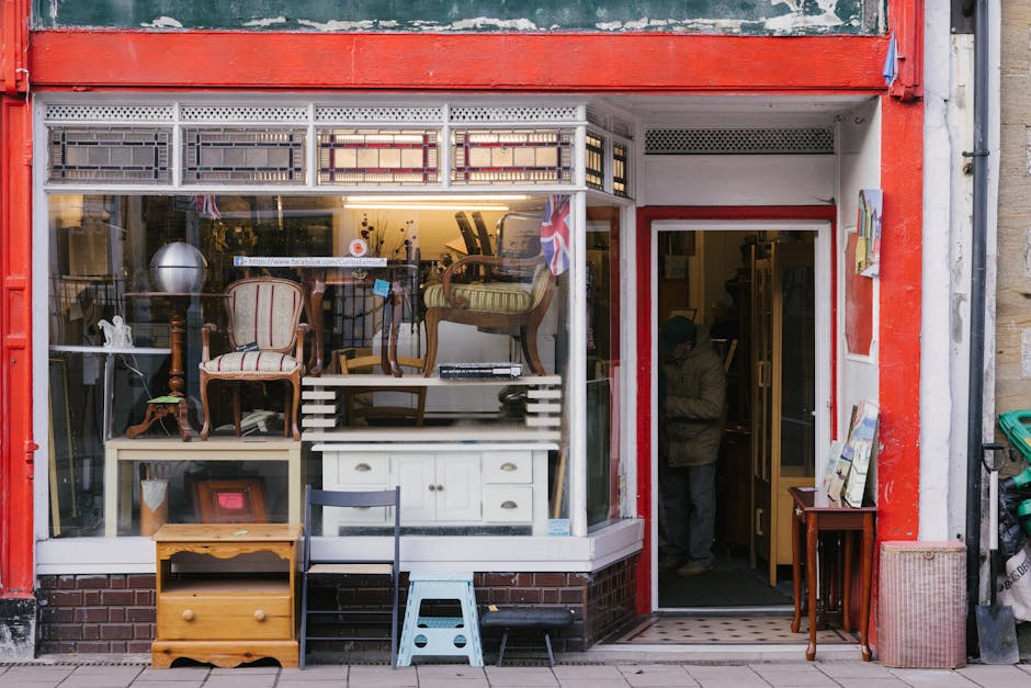 The image shows a busy street scene with various second-hand or discarded furniture and household items arranged along the pavement in front of a row of commercial buildings, including a pharmacy and a fish and seafood shop. There are sofas with visible fabric upholstery, tables, lamps, and other miscellaneous furniture items placed close together, some overlapping slightly. The furniture appears used, with some items showing signs of wear. Several people are gathered around the pile, some engaging in conversation or inspecting the items, while others stand nearby, possibly observing or waiting. A small scooter is parked to the left, along with a few other pedestrians in the background. The storefronts have signage, with one indicating a pharmacy and another a seafood business. The scene is lit by natural daylight, with a tree visible on the left side of the image, and the overall environment suggests an informal or on-site clearance situation, possibly related to local waste disposal or a community clearance, with Waste Removal Balham likely involved in the removal or collection of these items. The overall composition and context reflect typical roadside furniture and household item collection aimed at reuse or alternative waste handling.