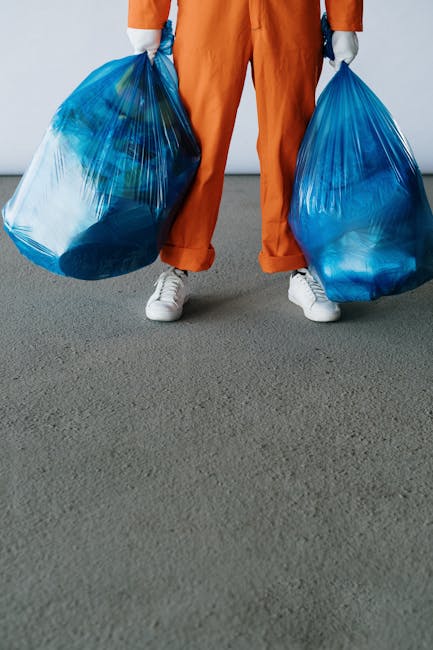 A person wearing orange work overalls and white sneakers is standing on a grey concrete surface, holding two large blue plastic rubbish bags filled with waste materials. The bags appear to contain household waste or collected debris, with their shiny, slightly translucent plastic showing the contents inside. The individual's hands are gloved, indicating handling of waste, and they are positioned against a plain, light-colored background. The scene reflects a typical setup for waste collection or rubbish removal, possibly as part of a private clean-up or alternative waste handling service, such as those offered by Waste Removal Balham. The focus is on the practical act of carrying refuse, emphasizing the aspect of rubbish collection that could be associated with various waste management options in an urban environment. The environment is indoor, with no specific environmental context beyond the concrete floor, and the lighting is neutral, highlighting the textures of the plastic bags and clothing. This scene visually illustrates the process of collecting waste items for disposal outside local authority bins, aligning with independent rubbish removal practices.
