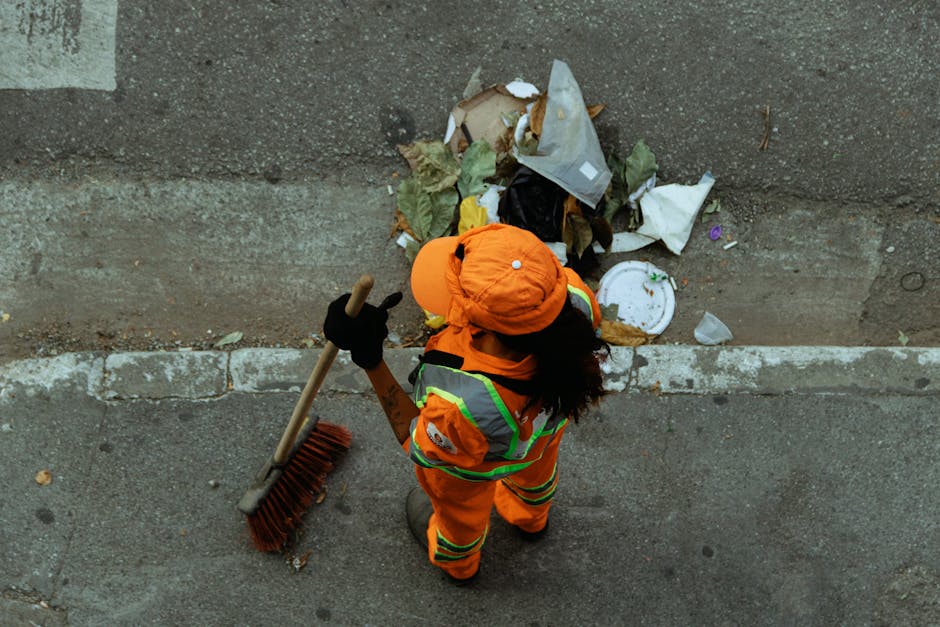 A person wearing orange work overalls and white sneakers is standing on a grey concrete surface, holding two large blue plastic rubbish bags filled with waste materials. The bags appear to contain household waste or collected debris, with their shiny, slightly translucent plastic showing the contents inside. The individual's hands are gloved, indicating handling of waste, and they are positioned against a plain, light-colored background. The scene reflects a typical setup for waste collection or rubbish removal, possibly as part of a private clean-up or alternative waste handling service, such as those offered by Waste Removal Balham. The focus is on the practical act of carrying refuse, emphasizing the aspect of rubbish collection that could be associated with various waste management options in an urban environment. The environment is indoor, with no specific environmental context beyond the concrete floor, and the lighting is neutral, highlighting the textures of the plastic bags and clothing. This scene visually illustrates the process of collecting waste items for disposal outside local authority bins, aligning with independent rubbish removal practices.