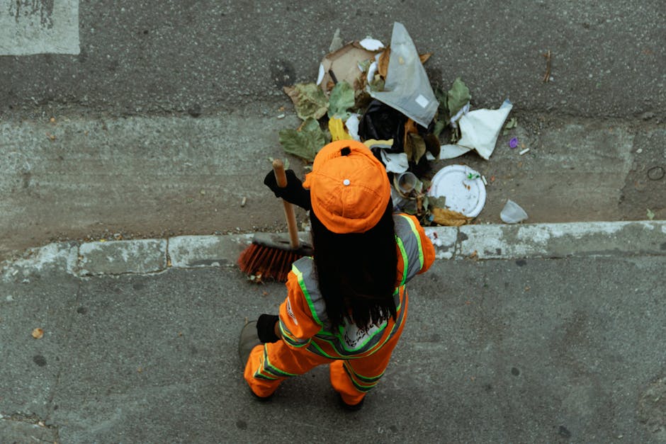 A person dressed in high-visibility orange work clothing and a matching orange cap is standing on a paved sidewalk, facing away from the camera. The individual is holding a broom with a wooden handle, actively sweeping a pile of mixed debris consisting of crumpled paper, dried leaves, and a white plastic plate with a green rim. The debris is located at the edge of a concrete curb, adjacent to a street surface made of asphalt. The environment appears to be a public urban area, possibly a roadside or sidewalk, with the focus on waste collection and rubbish clearing. The scene indicates an outdoor setting in daylight, with the person engaged in rubbish removal, representative of private waste management or on-site clearance services.