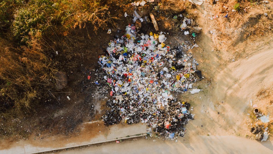 An aerial view of a small, open excavation site surrounded by dry, brown soil and sparse vegetation. In the center of the pit, there is a large pile of mixed waste materials, primarily composed of various plastic items such as bottles, bags, and packaging in different colours including white, blue, red, and black. The waste appears to be a combination of shredded plastic fragments and whole items, with some pieces partially buried or overlapping. Adjacent to the pile, the soil is exposed, showing a contrast between the lighter, compacted surface and the darker, looser material beneath. The scene suggests a private or alternative waste disposal activity, with the waste pile possibly awaiting removal or processing. Natural daylight illuminates the area, highlighting the textures and colours of the waste and the surrounding earth, consistent with an outdoor environment. Waste Removal Balham's services could be involved in the on-site clearance or removal of such waste materials, illustrating typical elements encountered in rubbish collection practices.